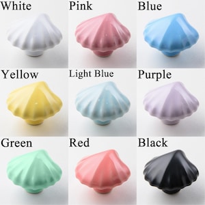 K&ouml;nnte beinhalten: Neun keramische Schrankkn&ouml;pfe in Muschelform in verschiedenen Farben: Wei&szlig;, Rosa, Blau, Gelb, Hellblau, Lila, Gr&uuml;n, Rot und Schwarz. Jeder Knopf hat eine gl&auml;nzende Oberfl&auml;che und einen kleinen Sockel.