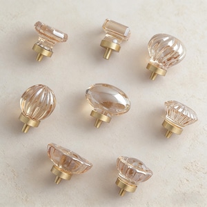 May include: A collection of eight decorative cabinet knobs. Each knob features a clear, amber-colored glass design atop a brass base. The knobs vary in shape, including rectangular, oval, and floral patterns, offering a range of aesthetic options.