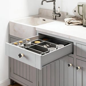 K&ouml;nnte beinhalten: Ein grauer K&uuml;chenschrank mit einer ge&ouml;ffneten Schublade, die eine organisierte Aufbewahrung zeigt. Die Schublade enth&auml;lt kleine Gl&auml;ser, Utensilien und Schalen. Der Schrank hat schwarze und goldfarbene Kleeblattkn&ouml;pfe. Ein wei&szlig;es Waschbecken und eine Armatur sind im Hintergrund zu sehen.