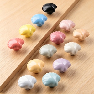 May include: A collection of seashell-shaped cabinet knobs in various colors, including black, blue, yellow, pink, and white. The knobs are made of ceramic and are arranged on a wooden surface. Each knob has a unique color and design.