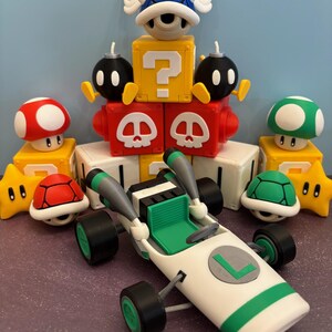 May include: A collection of colorful figurines inspired by a video game. The set includes a white and green race car, a blue shell with wings, mushroom and star figures, and blocks with question marks and skull designs.