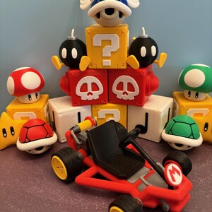 May include: A collection of colorful Mario-themed figurines and a red kart. The figurines include a red mushroom, a green mushroom, a star, a red turtle shell, a green turtle shell, a blue shell with wings, and black bomb characters.