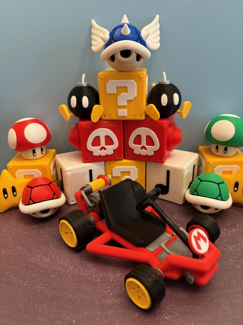 May include: A collection of colorful Mario-themed figurines and a red kart. The figurines include a red mushroom, a green mushroom, a star, a red turtle shell, a green turtle shell, a blue shell with wings, and black bomb characters.