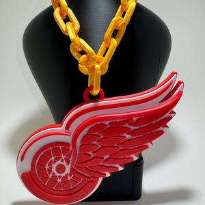 May include: A red and white Detroit Red Wings logo pendant on a gold chain necklace. The pendant is a 3D printed replica of the team's logo, featuring a wing with a red and white wheel inside.