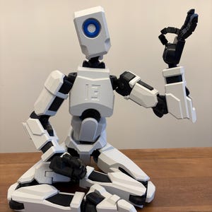 Puede incluir: Una figura de robot articulada en blanco y negro en posición sentada, con una mano levantada en un gesto. El robot tiene una cabeza cuadrada con un detalle de círculo azul y el número "13" en el pecho.