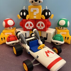 May include: A collection of colorful 3D-printed figures and a kart inspired by the Super Mario video game series. The kart is white with a red stripe and the letter "M". Other figures include a red mushroom, a green mushroom, and a blue shell.