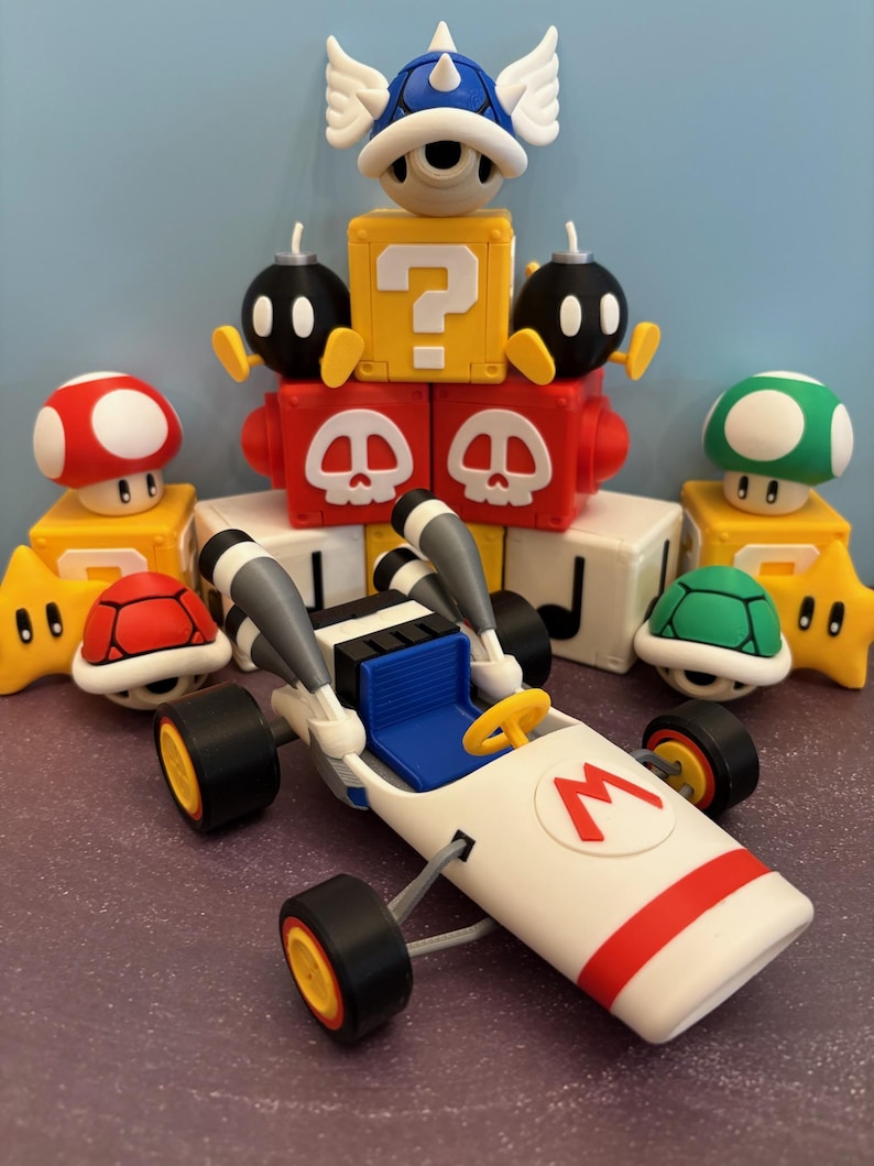 May include: A collection of colorful 3D-printed figures and a kart inspired by the Super Mario video game series. The kart is white with a red stripe and the letter "M". Other figures include a red mushroom, a green mushroom, and a blue shell.