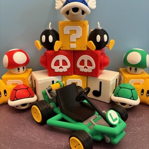 May include: A collection of colorful Mario-themed toys, including a green kart with yellow wheels, a blue shell with wings, red and green mushrooms, and various block figures. The toys are arranged on a purple surface.