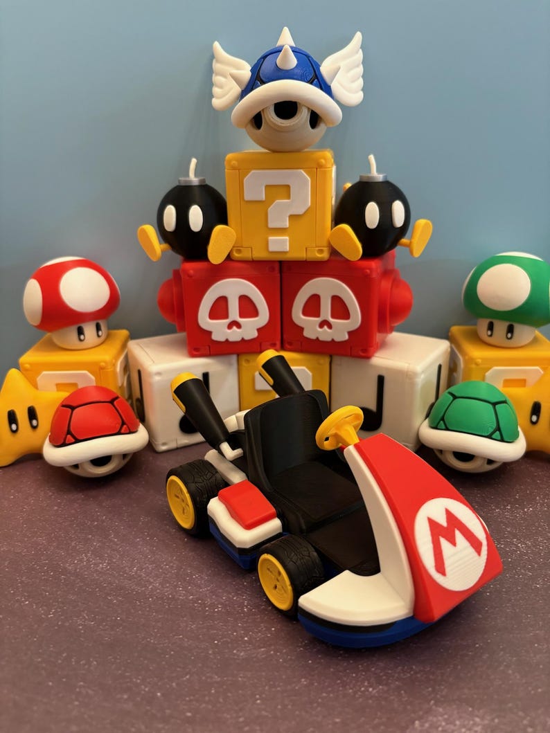 May include: A collection of colorful, 3D-printed figures inspired by the Mario Kart video game series. The scene includes a red and white kart, a blue shell, a question mark block, and various characters like mushrooms and turtles.