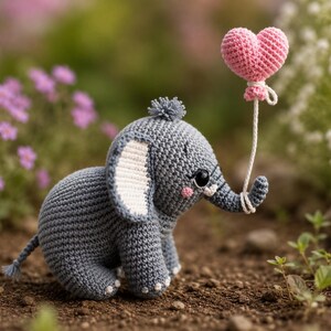 Könnte beinhalten: Ein graues gehäkeltes Elefanten-Spielzeug mit einem kleinen Haarbüschel auf dem Kopf, weißen Innenohren und rosa Wangen. Der Elefant hat ein schwarzes Auge und hält mit seinem Rüssel eine weiße Schnur, an der ein rosa gehäkelter Herzballon hängt.