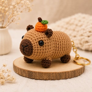 May include: A crocheted capybara keychain featuring a light brown body, dark brown snout, ears, and feet, with black bead eyes. A small orange fruit with a green leaf sits on top of the capybara's head. A gold-toned metal keyring is attached.
