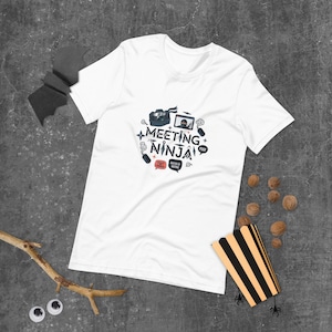 Könnte beinhalten: Weißes T-Shirt mit einem grafischen Design, das "Meeting Ninja" in einer kreisförmigen Anordnung zeigt. Das Design enthält Ninja-Symbole wie eine Kamera, Wurfsterne und Sprechblasen. Das Shirt ist aus weichem Material.