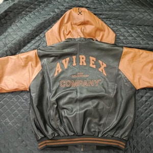 May include: A black and brown leather jacket with a hood. The jacket has brown sleeves and a brown hood. The back of the jacket features the word "AVIREX" in orange lettering, along with "NEW MILLENNIUM COMPANY". The jacket has a striped brown and orange band at the waist.