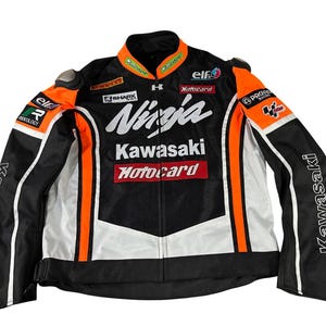 以下が含まれることがあります： 黒、白、オレンジのバイクジャケット。前面に「Ninja Kawasaki」と「Motocard」の文字がプリントされています。ジャケットはジッパーで閉められ、スタンドカラーです。