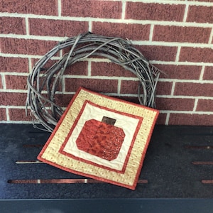 May include: A small, square quilted pumpkin wall hanging with a red and beige color scheme. The pumpkin is centered on a cream-colored background, framed by a red border and a beige patterned edge. A rustic twig wreath is behind the wall hanging.