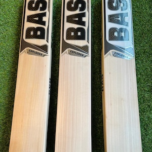 Puede incluir: Se muestran tres bates de críquet con hojas de madera clara y mangos grises. Los bates tienen la palabra "BAS" en letras negras grandes y en negrita en la parte superior, con "EXPLODER" impreso debajo. Están colocados sobre una superficie verde.