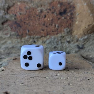 May include: Two white dice with black dots, one larger than the other, are displayed on a textured surface. The larger die shows five dots, while the smaller die shows one dot. The background is a brick wall.