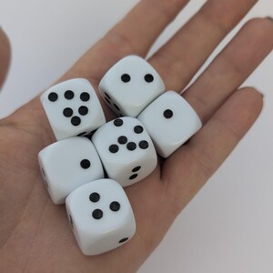 May include: Six white dice with black dots are held in a hand. The dice are arranged in a loose cluster, with each die displaying a different number of dots, from one to six. The dice are made of a smooth, glossy material.
