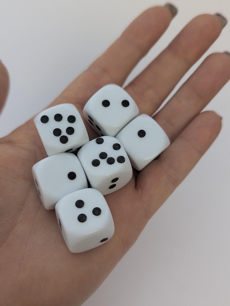 May include: Six white dice with black dots are held in a hand. The dice are arranged in a loose cluster, with each die displaying a different number of dots, from one to six. The dice are made of a smooth, glossy material.