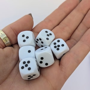 May include: Six white dice with black dots are held in a hand. The dice are standard six-sided cubes, each displaying a different number of dots. The dice are clean and appear new, suitable for various tabletop games.