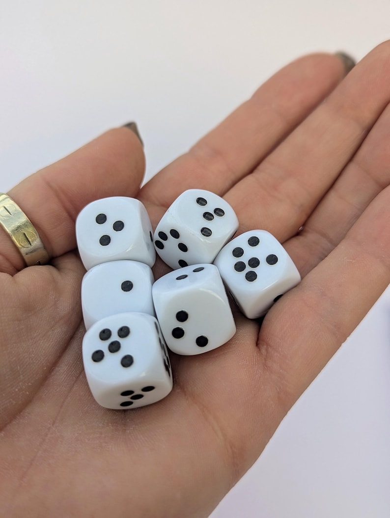 May include: Six white dice with black dots are held in a hand. The dice are standard six-sided cubes, each displaying a different number of dots. The dice are clean and appear new, suitable for various tabletop games.