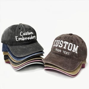 May include: A stack of baseball caps in various colors, including black, brown, and blue. The caps have a distressed look and feature the text "Custom Embroidery" and "CUSTOM YOUR TEXT" in white lettering. The caps are made of a soft, worn material.