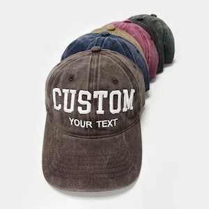 May include: A selection of baseball caps in shades of brown, navy, and burgundy. The front cap is brown with the white embroidered text "CUSTOM" and "YOUR TEXT". The caps are arranged in a diagonal line, showcasing their different colours.