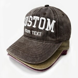 May include: A stack of three baseball caps in shades of brown, olive green, and purple. The top cap is a distressed brown with the word "CUSTOM" and "YOUR TEXT" embroidered in white. The caps are stacked on a white background.