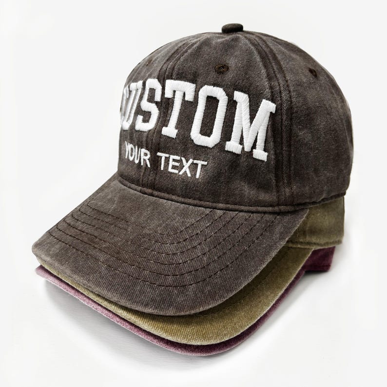 May include: A stack of three baseball caps in shades of brown, olive green, and purple. The top cap is a distressed brown with the word "CUSTOM" and "YOUR TEXT" embroidered in white. The caps are stacked on a white background.
