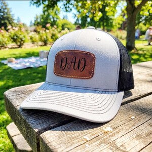 May include: A gray and white trucker hat with a black mesh back. The hat features a brown leather patch with the word "DAD" in cursive script. The hat is sitting on a wooden picnic table.