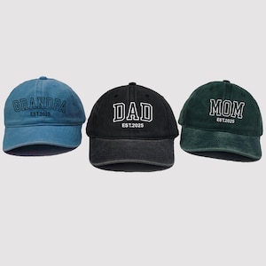 May include: Three baseball caps in blue, black, and green. Each cap has a different word in white block letters: "GRANDPA", "DAD", and "MOM". Below each word is "EST.2025". The caps are made of a soft material.