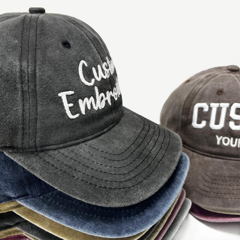 May include: A stack of baseball caps in various colors, including gray, blue, and brown. The front cap is gray with the words "Custom Embroidered" in white. The brown cap has the word "CUS" embroidered in white.
