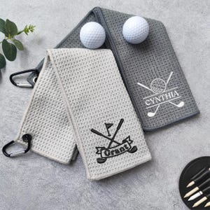 May include: Two golf towels, one light grey and one dark grey, with a waffle weave texture. The light grey towel has the name "Grant" embroidered. The dark grey towel has the name "Cynthia" embroidered. Two golf balls are on top.