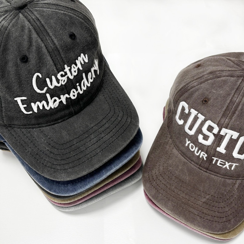 May include: A stack of custom embroidered baseball caps in various colors, including black, blue, and brown. The caps have curved visors and adjustable straps. The text "Custom Embroidery" and "Your Text" are embroidered in white on the front of the caps.