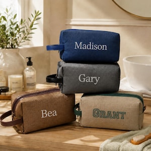 May include: Four rectangular toiletry bags with side handles, each featuring a personalised name embroidered on the front. The bags are in navy blue with Madison, dark grey with Gary, brown with Bea, and light tan with the green text GRANT.