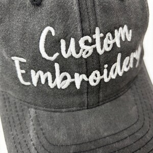 May include: A charcoal gray baseball cap with the words "Custom Embroidery" embroidered in white script across the front. The cap has a curved brim and a button on top.