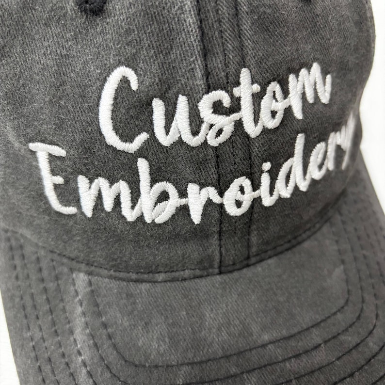 May include: A charcoal gray baseball cap with the words "Custom Embroidery" embroidered in white script across the front. The cap has a curved brim and a button on top.