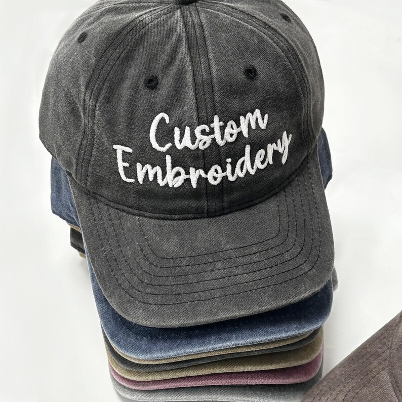 May include: A stack of gray, blue, and brown baseball caps. The top cap is gray with the words "Custom Embroidery" embroidered in white. The caps have a classic, curved brim and adjustable strap.