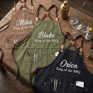 May include: Three canvas aprons in brown, olive green, and black, each featuring cross-back straps with leather accents, front pockets, and white text reading Miles, Blake, or Orion followed by the phrase King of the BBQ in a script and sans-serif font.