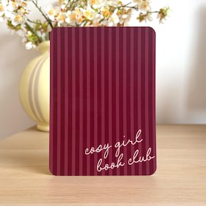 May include: A rectangular card with a deep burgundy background featuring vertical stripes in a slightly darker shade. The text "cosy girl book club" is written in a white, cursive, handwritten-style font in the lower right corner.
