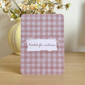 May include: A rectangular card featuring a dusty pink and white gingham pattern. In the centre, a white paper scrap with torn edges is attached with tape, displaying the dark brown cursive text "booked for autumn" on its surface.