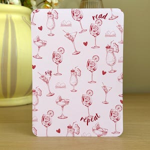 May include: A rectangular card with a light pink background featuring a repeating pattern of red line-art illustrations of various cocktails, small open books, and hearts. The text "read", "sip", and "repeat" is printed in red script.
