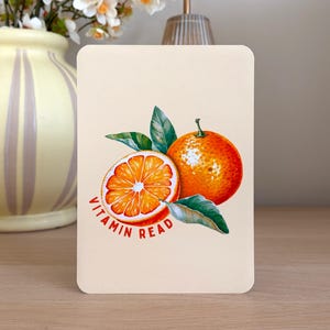 Könnte beinhalten: Eine rechteckige, cremefarbene Karte mit einer lebendigen Illustration einer ganzen Orange und einer aufgeschnittenen Orangenhälfte mit grünen Blättern. Der Text VITAMIN READ ist in roten Großbuchstaben in einem Bogen unter der Frucht gedruckt.