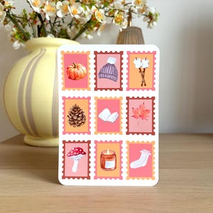 May include: A rectangular sticker sheet featuring nine individual stamp-shaped designs arranged in a grid. The illustrations include a pumpkin, a knit beanie, marshmallows on sticks, a pinecone, an open book, a maple leaf, a mushroom, a candle, and a sock.