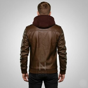 May include: Brown leather jacket with a burgundy hood, featuring a ribbed hem and cuffs. The jacket has a classic design with shoulder accents and a front zipper. The back view showcases the jacket's tailored fit and the "NOBLE JACKETS" logo.