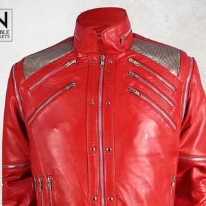 Puede incluir: Chaqueta de cuero roja vibrante con cremalleras plateadas y detalles con tachuelas. La chaqueta presenta un cuello alto y hombreras plateadas. El logotipo de la marca "NOBLE JACKETS" es visible en la esquina superior izquierda.