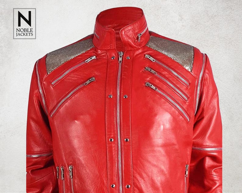 Puede incluir: Chaqueta de cuero roja vibrante con cremalleras plateadas y detalles con tachuelas. La chaqueta presenta un cuello alto y hombreras plateadas. El logotipo de la marca "NOBLE JACKETS" es visible en la esquina superior izquierda.