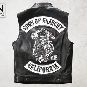 May include: Black leather vest with the text "SONS OF ANARCHY" and "CALIFORNIA" in white lettering. The back of the vest features a skull graphic with a scythe and rifle. The vest has a button closure and the brand "NOBLE JACKETS" is visible.