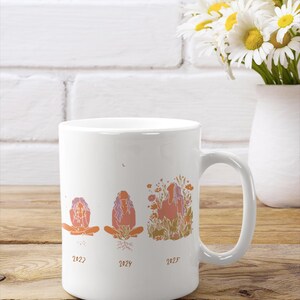 May include: White ceramic mug featuring a graphic of three figures in different stages, with the years 2022, 2024, and 2025 below each. The figures are in shades of orange and purple. A bouquet of daisies in a white vase is in the background.
