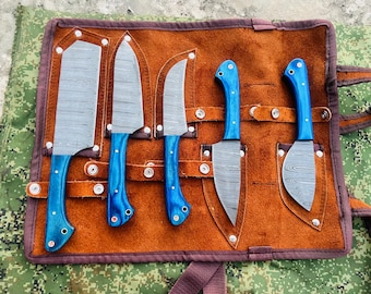 May include: A set of six kitchen knives with blue wooden handles and patterned steel blades, displayed in a brown leather roll. The knives vary in size and shape, with a cleaver and paring knives included. The roll has a brown exterior and a dark brown interior.